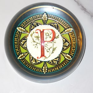 May include: A round, decorative paperweight with a dark teal and gold border. The center features a white circle with a red letter "P" and floral accents. The design is surrounded by a green and gold pattern.