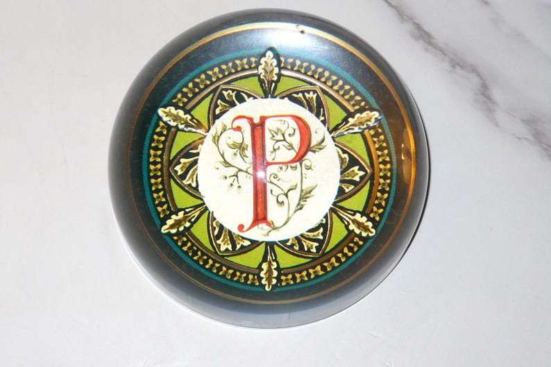May include: A round, decorative paperweight with a dark teal and gold border. The center features a white circle with a red letter "P" and floral accents. The design is surrounded by a green and gold pattern.