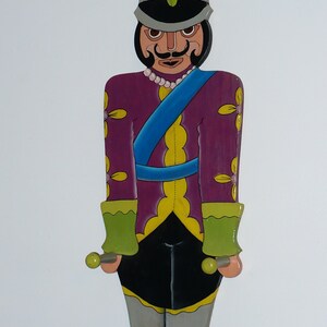 May include: A flat, wooden nutcracker wall decoration featuring a purple jacket with yellow floral accents, a blue sash, and a black hat with a green feather. The figure has a black mustache, a white pearl-like necklace, green cuffs, and grey pants.