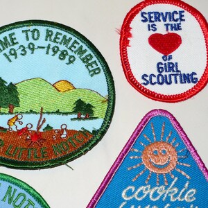 May include: A collection of vintage Girl Scout patches. One patch reads "A Time to Remember 1939-1989" with a landscape scene. Another says "Service is the of Girl Scouting" with a red heart. A triangular patch features a smiling sun.