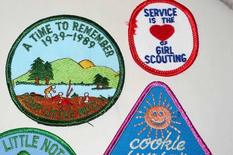 May include: A collection of vintage Girl Scout patches. One patch reads "A Time to Remember 1939-1989" with a landscape scene. Another says "Service is the of Girl Scouting" with a red heart. A triangular patch features a smiling sun.