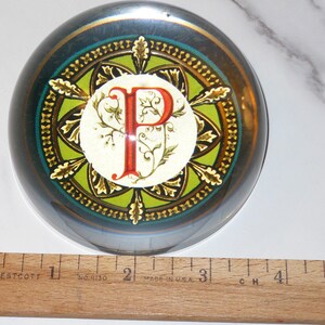 May include: A round, glass paperweight with a decorative design. The paperweight features a central design with the letter "P" in red, surrounded by a green and gold pattern. A wooden ruler is placed next to the paperweight, measuring up to 5 inches.
