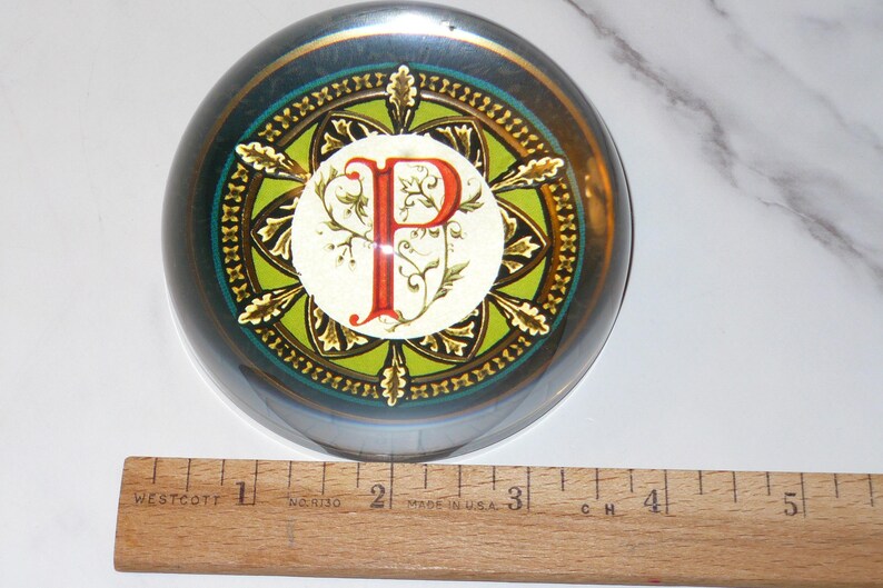 May include: A round, glass paperweight with a decorative design. The paperweight features a central design with the letter "P" in red, surrounded by a green and gold pattern. A wooden ruler is placed next to the paperweight, measuring up to 5 inches.