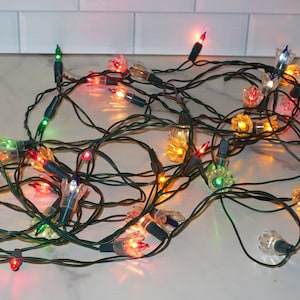 May include: A string of decorative holiday lights featuring dark green wiring and clear, flower-shaped plastic covers over each bulb. The lights emit a variety of colors including red, green, blue, orange, and yellow, creating a multi-colored glow.