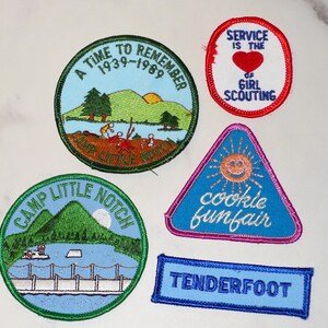 May include: A collection of embroidered patches with various designs and text. The patches include a round patch with the text "A TIME TO REMEMBER 1939-1989", a triangular patch with a smiling sun and the words "cookie funfair", and a rectangular patch that says "TENDERFOOT".
