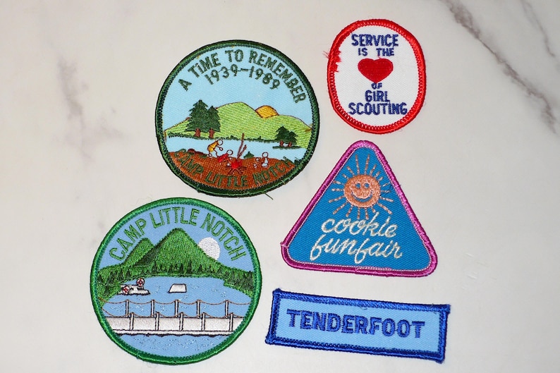 May include: A collection of embroidered patches with various designs and text. The patches include a round patch with the text "A TIME TO REMEMBER 1939-1989", a triangular patch with a smiling sun and the words "cookie funfair", and a rectangular patch that says "TENDERFOOT".