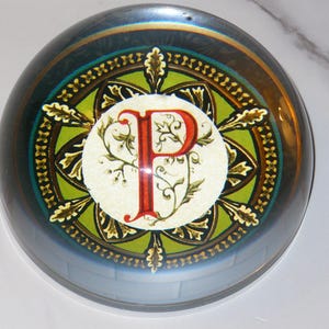May include: A round, glass paperweight with a decorative design. The design features a large red letter "P" in the center, surrounded by a green and gold pattern. The paperweight has a reflective, iridescent finish.