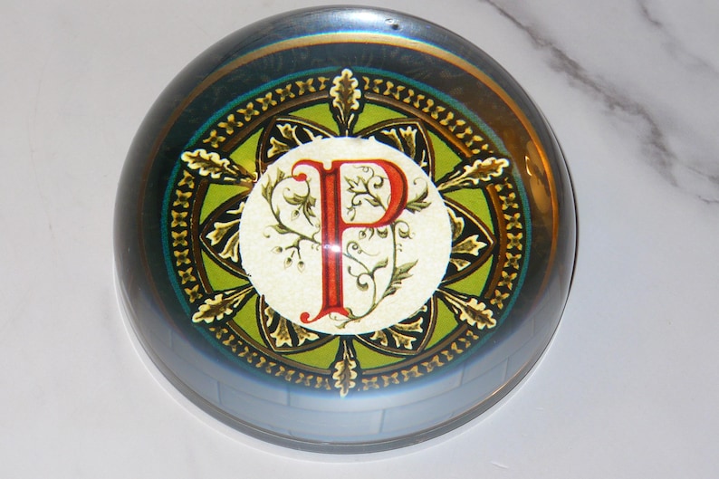 May include: A round, glass paperweight with a decorative design. The design features a large red letter "P" in the center, surrounded by a green and gold pattern. The paperweight has a reflective, iridescent finish.