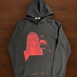 May include: Dark gray hooded sweatshirt with a red graphic of a woman and a shark. The sweatshirt has a front pocket and a drawstring hood. The graphic is a bold, eye-catching design.