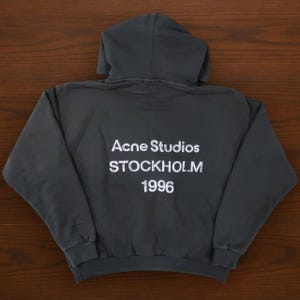 May include: Dark gray hoodie with a hood and long sleeves. The back of the hoodie features the white text "Acne Studios STOCKHOLM 1996". The hoodie is made of a soft material.