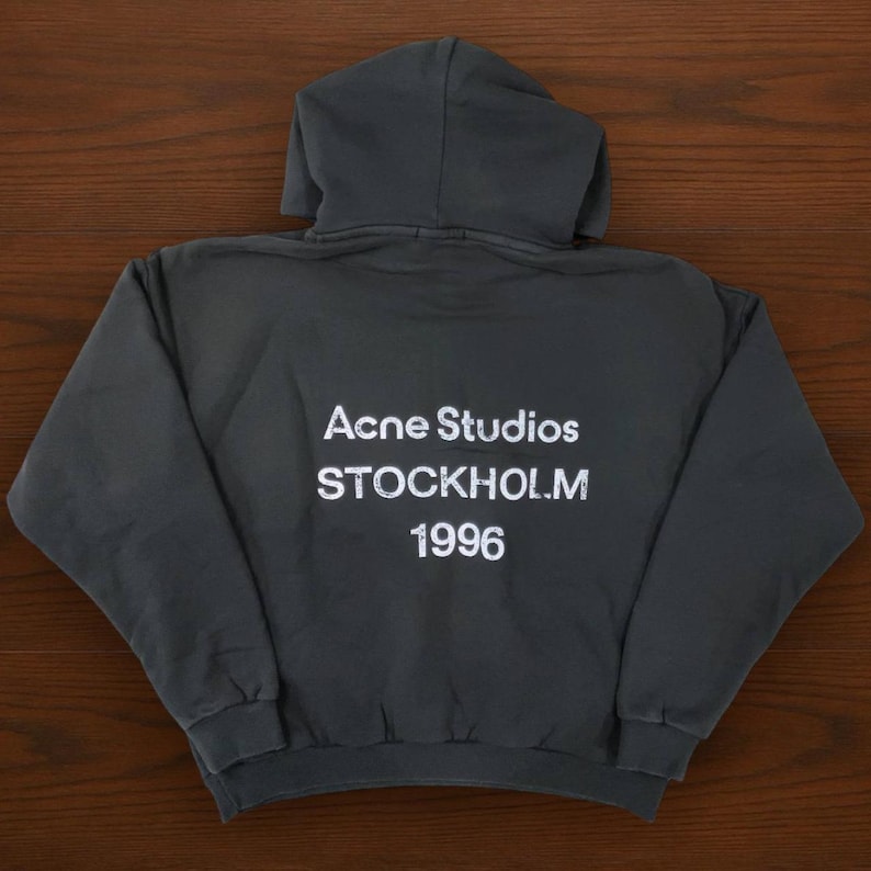 May include: Dark gray hoodie with a hood and long sleeves. The back of the hoodie features the white text "Acne Studios STOCKHOLM 1996". The hoodie is made of a soft material.