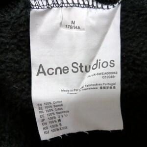 May include: A close-up of a white clothing tag with the text "Acne Studios" in gray. The tag also includes size information, fabric composition, and the text "Made in Portugal". The background is black.