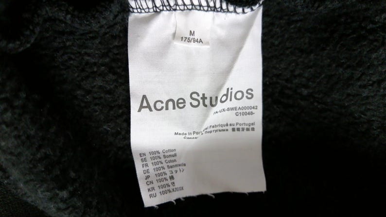 May include: A close-up of a white clothing tag with the text "Acne Studios" in gray. The tag also includes size information, fabric composition, and the text "Made in Portugal". The background is black.
