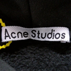 May include: Close-up of a black hooded sweatshirt with a white label. The label reads "Acne Studios" in black text. A yellow plastic tag is attached to the label.