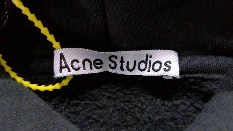 May include: Close-up of a black hooded sweatshirt with a white label. The label reads "Acne Studios" in black text. A yellow plastic tag is attached to the label.