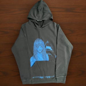 May include: Dark gray hoodie with a front pocket and a hood. The front features a blue graphic of a woman with long hair and a shark. The text at the bottom of the hoodie is blurry.