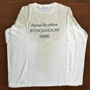 以下が含まれることがあります： 黒字で「Acne Studios STOCKHOLM 1996」と書かれた白い長袖Tシャツ。Tシャツは柔らかい素材でできており、クルーネックです。木製の表面に平らに置かれたTシャツ。