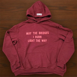 以下が含まれることがあります： フロントポケットとフードが付いた、マルーン色のパーカー。「MAY THE BRIDGES I BURN LIGHT THE WAY」の文字が、前面に明るい赤色でプリントされています。ブランド名「VETEMENTS」がフードにプリントされています。