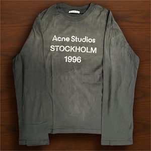 May include: An olive-green long-sleeved shirt featuring the white text "Acne Studios STOCKHOLM 1996". The shirt has a crew neck and a faded look. The garment appears to be made of a soft material.