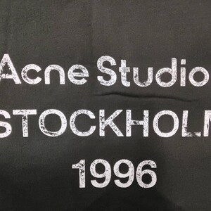 May include: Black fabric with the white text "Acne Studios STOCKHOLM 1996". The text has a distressed, vintage appearance. The letters are bold and easily readable against the dark background.