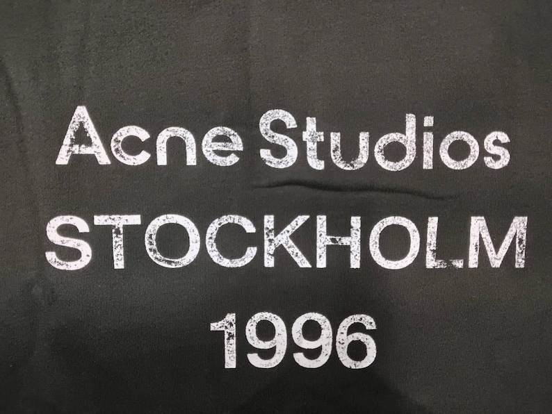May include: Black fabric with the white text "Acne Studios STOCKHOLM 1996". The text has a distressed, vintage appearance. The letters are bold and easily readable against the dark background.