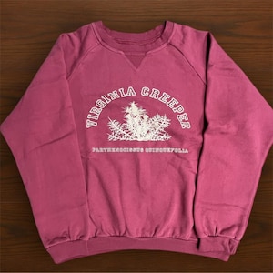 May include: A maroon sweatshirt with the words "VIRGINIA CREEPER" in a white arch above a white illustration of a plant. Below the illustration is the text "PARTHENOCISSUS QUINQUEFOLIA". The sweatshirt has a crew neck and long sleeves.