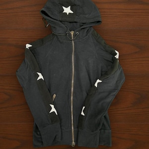 May include: A black zip-up hoodie featuring a white star printed on the hood and a series of white stars running down the length of each sleeve. The garment includes a full-length metal zipper, a drawstring hood, and two front pockets.