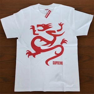 May include: White t-shirt featuring a red dragon graphic and the word "SUPREME" in red. The shirt has a tag that says "SUPREME" in red and white. The shirt is folded on a brown surface.