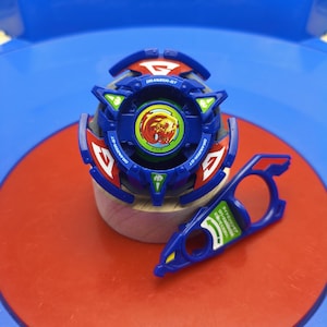 May include: A blue and red spinning top toy featuring the text Dranzer-GT, with a central circular emblem depicting a stylized red phoenix. Beside it lies a blue plastic ripcord tool with a green sticker containing Japanese text and an arrow.