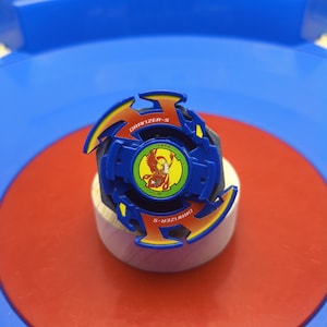 May include: A blue and orange plastic spinning top toy featuring a central circular emblem with a yellow phoenix graphic. The top is marked with the text DRANZER-S and DRZ2, and it includes small yellow accents on the outer edges of the structure.