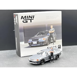 May include: A miniature silver Nissan Skyline GT-R (R32) model car with a matching figure. The box behind it features the car and figure, with the words "MINI GT" and Japanese text. The car has black wheels.