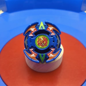 May include: A blue and orange spinning top toy featuring a central circular emblem with a red phoenix graphic and green border. The top has four symmetrical arms with white dragon motifs and green triangular accents, with text reading Dranzer V2.