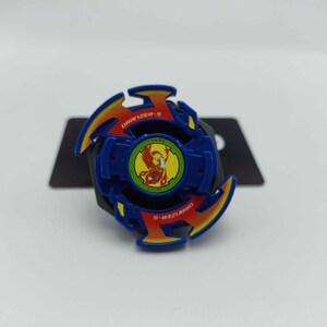 Pu&ograve; includere: Un giocattolo Beyblade Dranzer-S blu, giallo e rosso. Il giocattolo ha un design circolare con un emblema centrale e la scritta "DRANZER-S" sull'anello esterno. Il giocattolo &egrave; su uno sfondo nero.