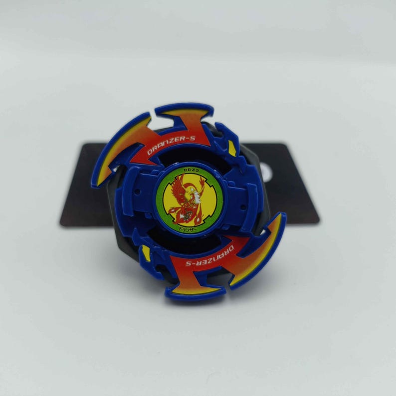 Pu&ograve; includere: Un giocattolo Beyblade Dranzer-S blu, giallo e rosso. Il giocattolo ha un design circolare con un emblema centrale e la scritta "DRANZER-S" sull'anello esterno. Il giocattolo &egrave; su uno sfondo nero.