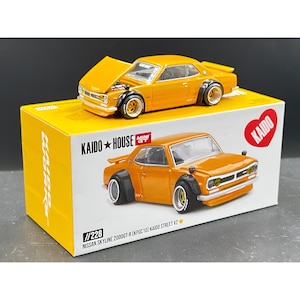 May include: A diecast model of a Nissan Skyline 2000GT-R (KPGC10) Kaido Street V2 in orange. The model car is on top of a yellow and white box with the text "KAIDO HOUSE" and "MINI GT". The car's hood is open.