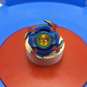 May include: A blue and black spinning top toy featuring four orange and yellow wing-like protrusions. The center circular emblem displays a red phoenix graphic on a green background with the text DRANZER written in small black letters.