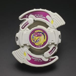 May include: A white and purple Beyblade Seaborg toy with a circular design. The toy features the word "DEFENSE" and "BEYBLADE SEABORG" printed on it. The centre of the toy has a circular graphic with a creature design and Japanese text.
