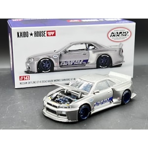 Könnte beinhalten: Ein silbernes Nissan Skyline GT-R (R34) Modellauto mit geöffneter Motorhaube, das Motordetails zeigt. Das Auto hat blaue Räder und ein "KAIDO HOUSE"-Logo. Die Verpackung zeigt ein ähnliches Auto. Der Text "KAIDO HOUSE MINI GT" ist sichtbar.