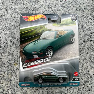Puede incluir: Un coche de juguete Hot Wheels Premium de un Mazda MX-5 Miata '91 verde. El embalaje muestra el coche en una carretera, con el texto "Modern Classics" y "Real Riders". El coche es a escala 1:64, de unos 6.35 cm de largo.