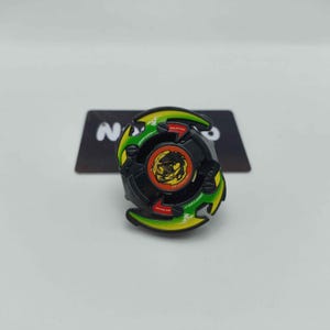 May include: A black, green, and yellow spinning top toy with a dragon design at its centre. The toy is circular with blades and a red and yellow central design. It rests on a black card with the letter "N" visible.