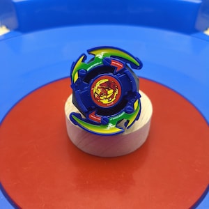 May include: A blue and green spinning top toy featuring a central circular emblem with a red phoenix graphic. The toy has curved, wing-like edges with white text that reads DRANZER-F and includes small green dragon icons on the outer frame.