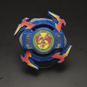 May include: A blue, red, and yellow toy with a circular design. The toy has a central emblem with a red bird and Japanese text. The toy has three red and yellow wing-shaped protrusions. The toy is on a dark surface.