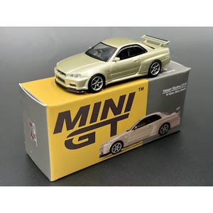 Op de afbeelding: Een goudkleurig modelauto, een Nissan Skyline GT-R, staat op zijn doos. De doos is geel en grijs met de woorden "MINI GT" in grote, vette letters. De doos bevat ook een afbeelding van de auto.