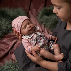 Puede incluir: Una muñeca bebé realista con gorro de punto rosa, cárdigan y conjunto con estampado floral. La muñeca está en brazos de alguien, con los ojos cerrados. La piel de la muñeca tiene un tono realista.