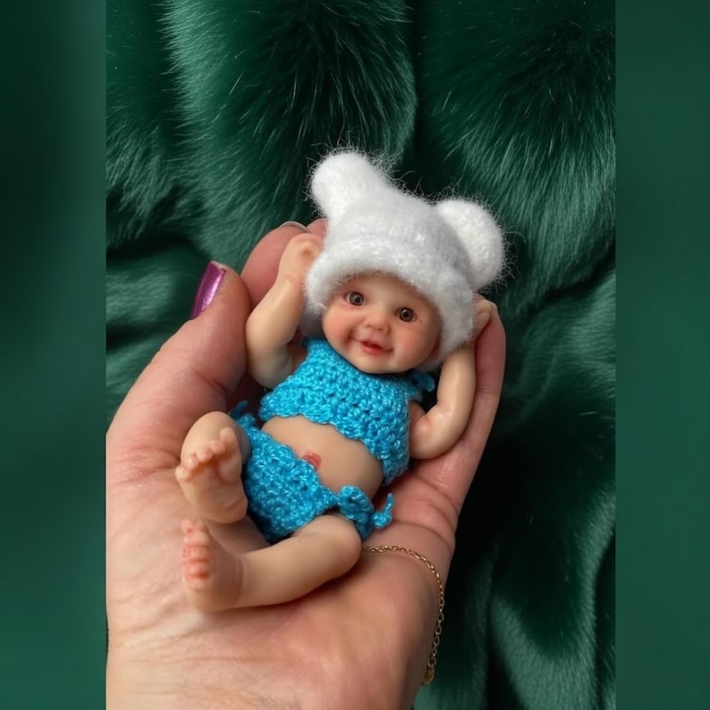 May include: A small baby doll dressed in a blue crocheted outfit and a white hat with bear ears. The doll has a smiling face and is held in an open hand. The background is a dark green, textured fabric.
