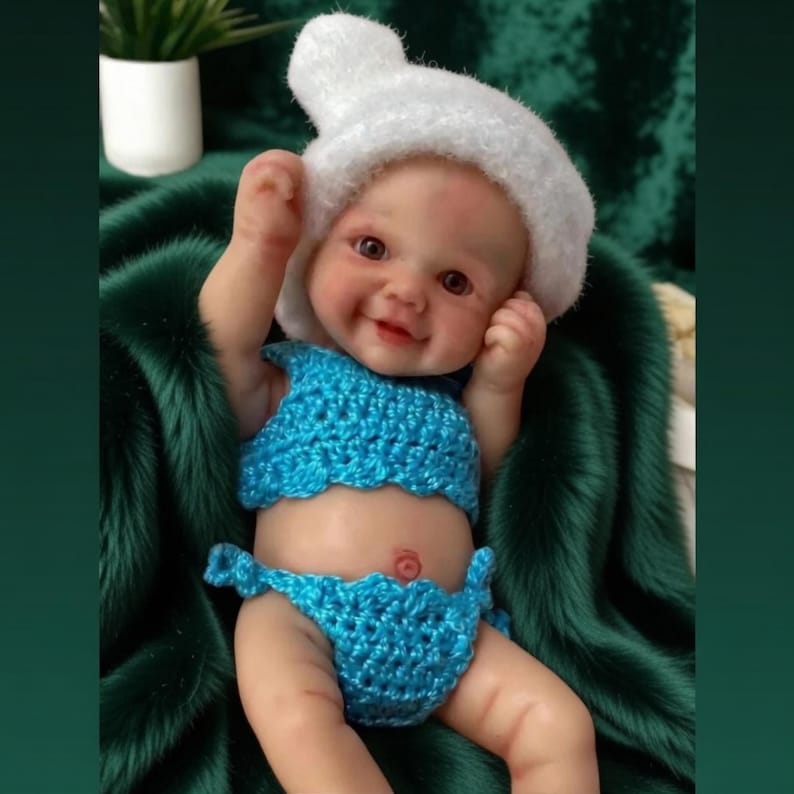 Op de afbeelding: Een levensechte babypop met een gehaakte blauwe bikini en een witte hoed. De pop lacht en is geplaatst op een donkergroen, getextureerd oppervlak. De pop heeft een lichte huid en gedetailleerde gelaatstrekken.