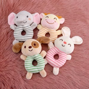 May include: Five plush baby rattles in the shape of animals: an elephant, a cow, a dog, and a rabbit. Each has a striped ring body in various colors. The elephant is gray with pink ears, the cow is yellow with brown horns, the dog is tan with brown ears, and the rabbit is white.