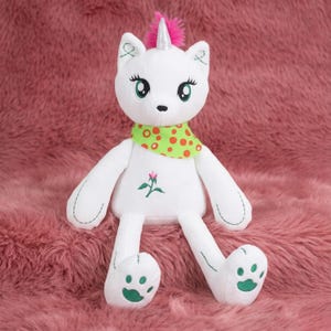May include: A white plush cat-unicorn hybrid toy with a pink horn and mane. It has green eyes, a green and orange polka-dot scarf, and a flower embroidered on its chest. The paws have green paw prints.