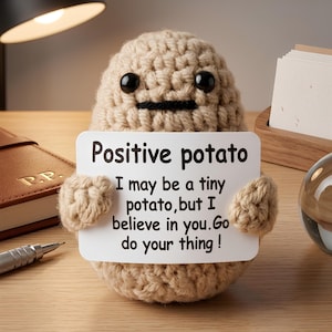 Könnte beinhalten: Eine beige gehäkelte Kartoffelfigur mit schwarzen Perlenaugen und einem kleinen schwarzen gestickten Mund. Sie hält ein weißes rechteckiges Schild mit der Aufschrift: Positive potato, I may be a tiny potato, but I believe in you. Go do your thing!