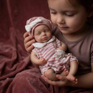 Op de afbeelding: Een levensechte babypop gekleed in een roze en wit gestreept outfit en bijpassende hoed met een witte strik. De pop wordt door een persoon vastgehouden. De ogen van de pop zijn gesloten en het lijkt te slapen. De outfit van de pop heeft een witte kraag en kanten rand.